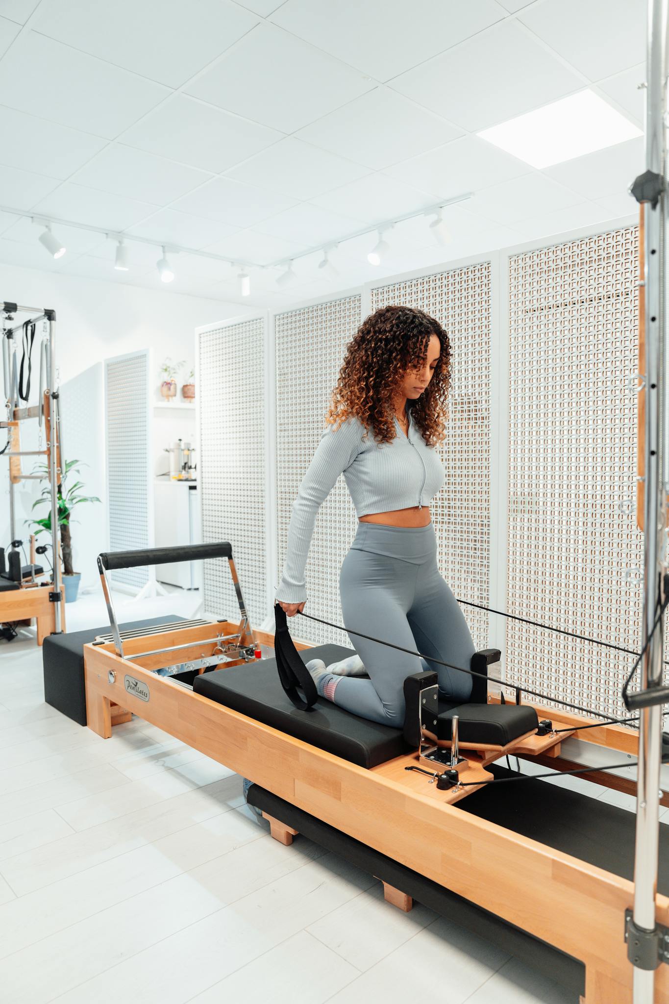 Woman in gray tracksuit exercises on Pilates tower in a bright white studio, showcasing fitness and concentration.